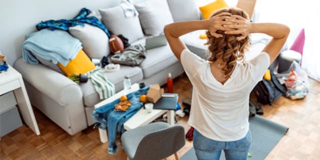 Lady standing with her arms behind her head with her back to the camera facing clutter on her floor at sofa in the background.