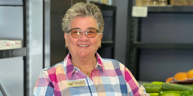 photo of a volunteer at pie in the sky smiling, standing next to fresh fruit and vegetables in a warehouse.