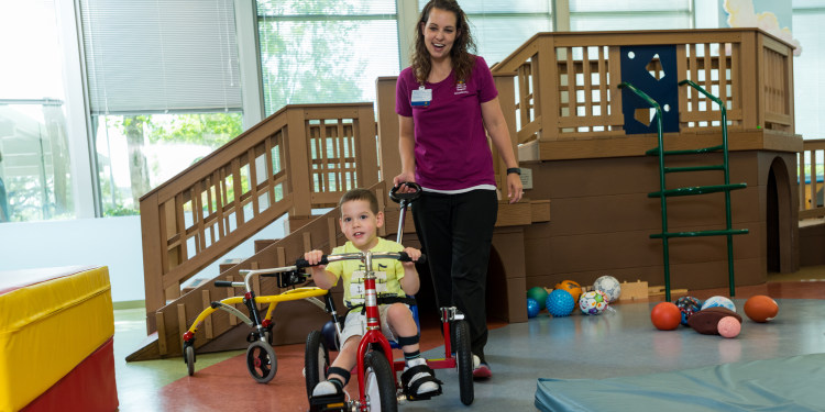 photo of a pediatric clinic setting where a female provider pushes a young male patient on a tricycle