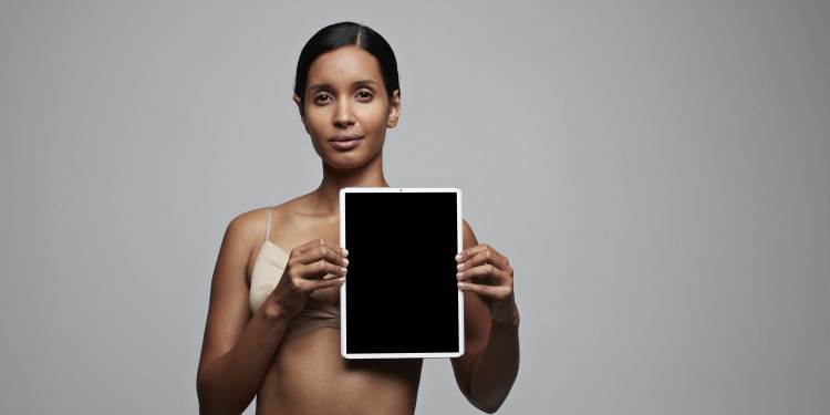 woman in underwear standing against a neutral background and holding a screen over her left breast