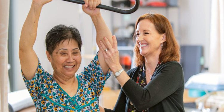 photo of a adult rehab clinic setting where a female therapist helps a middle-aged Asian woman perform arm exercises
