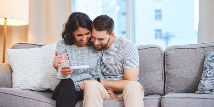 A happy young couple sitting on the sofa together and holding a positive pregnancy test
