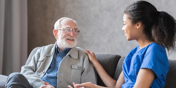 A caring black nurse talks to an elderly senior male patient sitting in the living room at a homecare visit