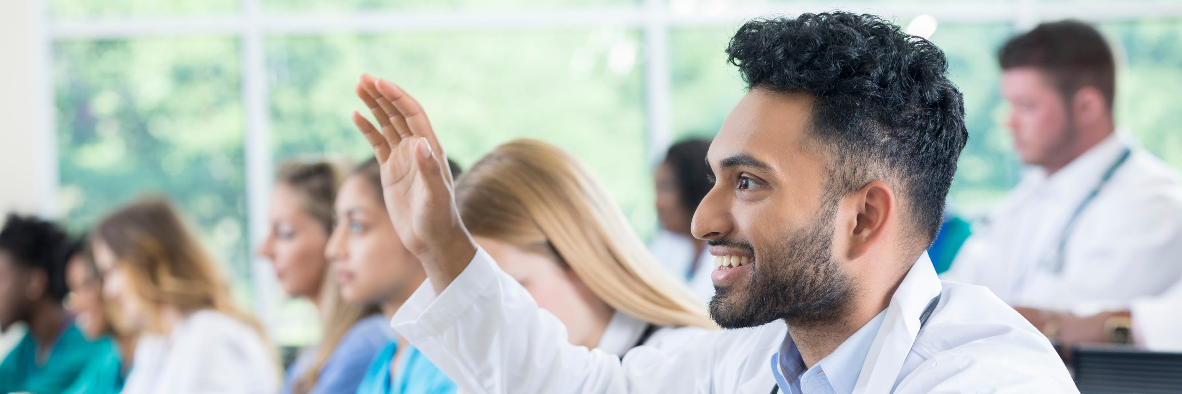 young male doctor raising his hand in a classroom setting with colleagues in the background