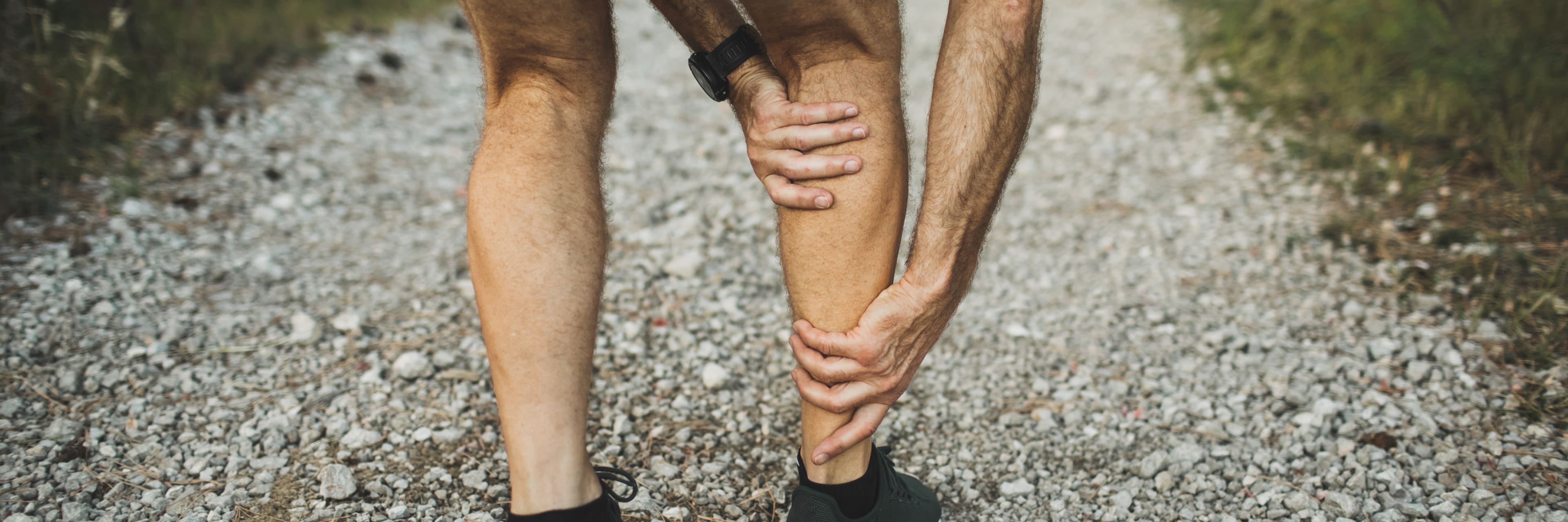 view of a man from behind wearing shorts and athletic shoes that was exercising on a gravel walking path is bent over holding his right calf from pain.
