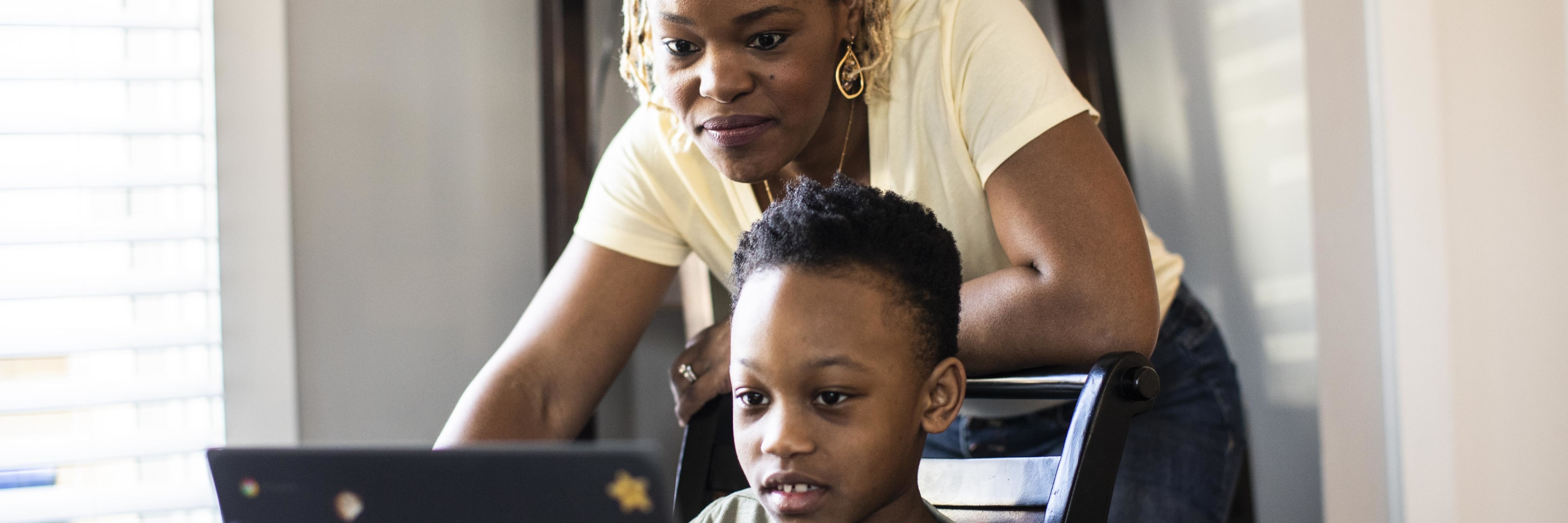 an African American mother looks over her son's shoulder as they both look at the screen of a laptop computer.