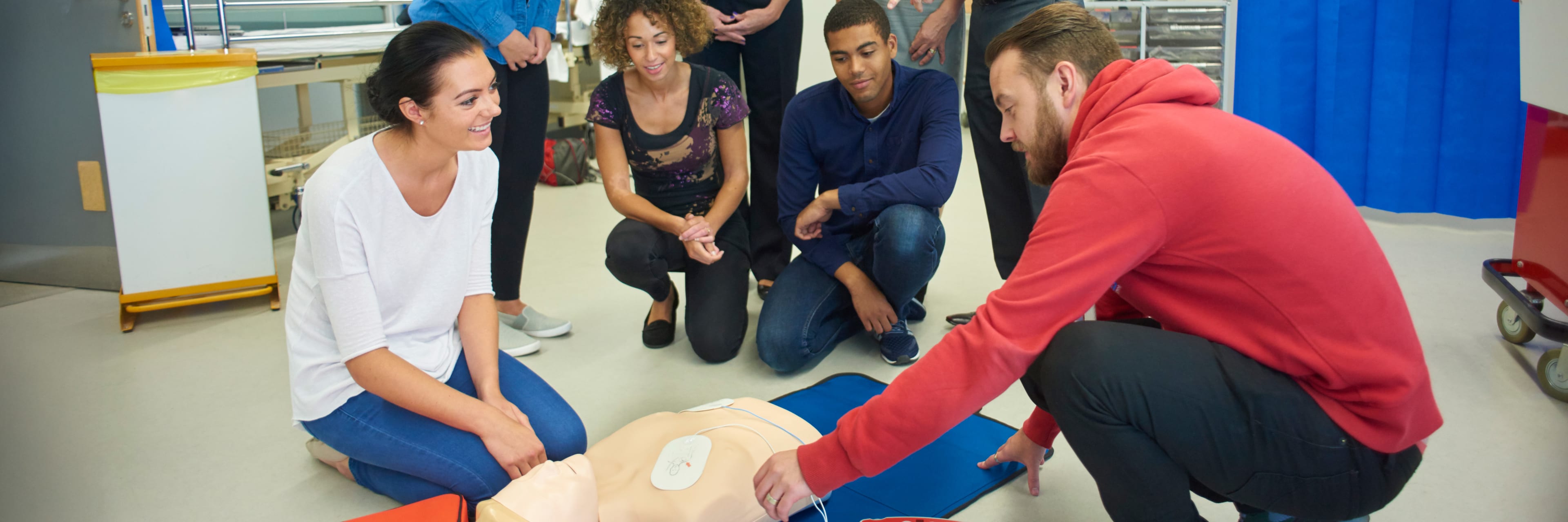 a mixed age group listen to teacher as he shows the procedure involved to resuscitate using a defibrillator