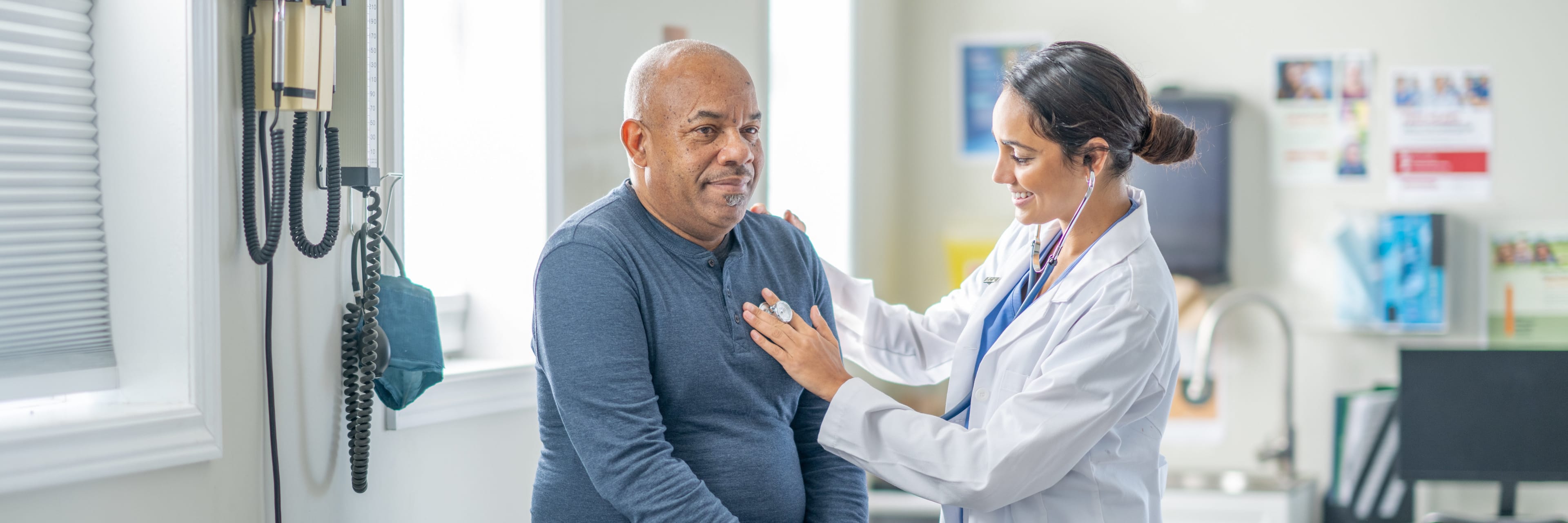 a female cardiologist places a stethoscope over a male patient's heart on an exam table in an office setting.