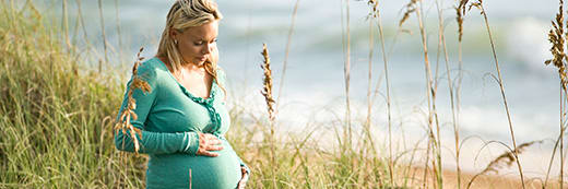 a pregnant woman looks down at her baby bump while she stands in the midst of sand dunes at the oceanfront.