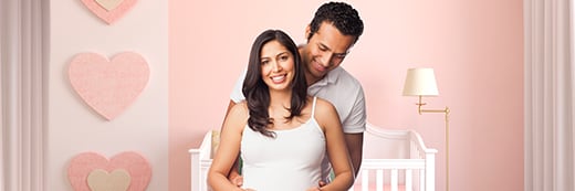 A male/female couple holds a picture of a sonogram while standing in a pink nursery