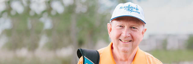 photo of patient Jim Marshall wearing an orange shirt and baseball cap standing on a golf course and carrying a golf bag on his shoulder