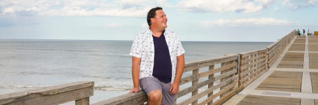 slightly smiling man leaning on a boardwalk at the beach