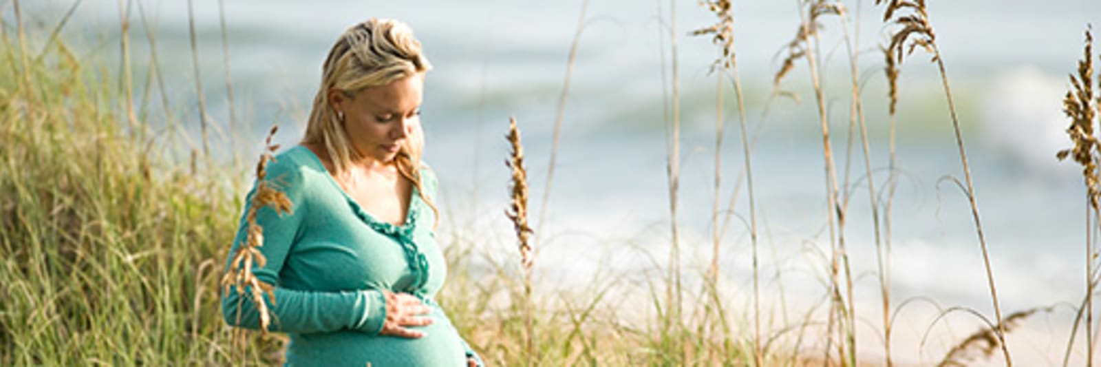 a pregnant woman looks down at her baby bump while she stands in the midst of sand dunes at the oceanfront.