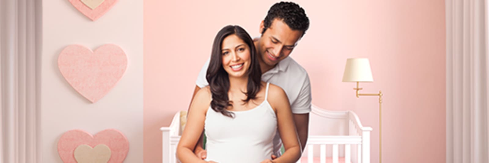 A male/female couple holds a picture of a sonogram while standing in a pink nursery