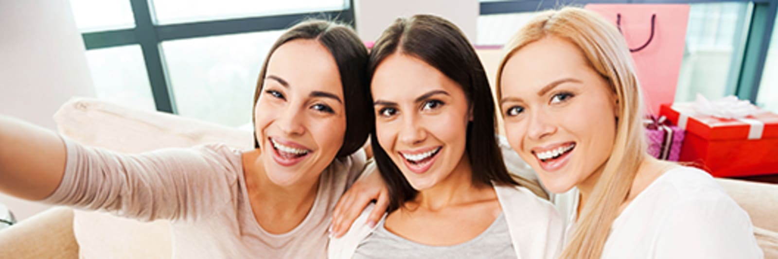 happy young pregnant woman holding baby booties on her abdomen and smiling while two friends sitting close to her and making selfie