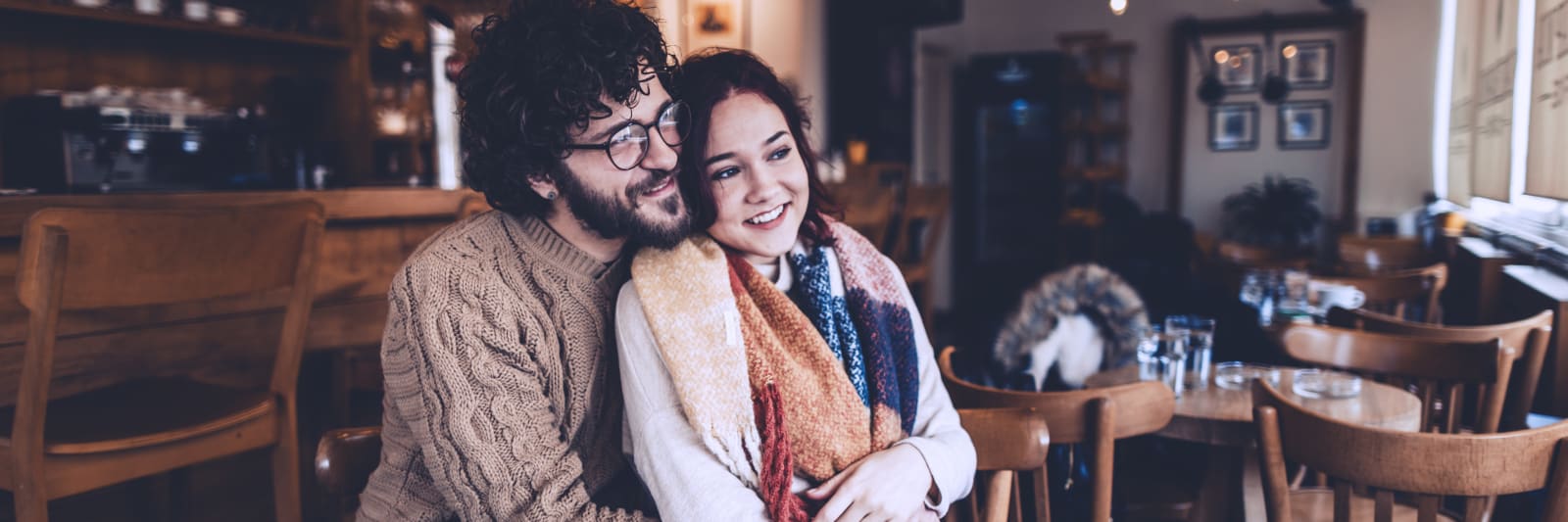 Young couple sitting at a table in a coffee shop smiling as they look out the window while cuddling.