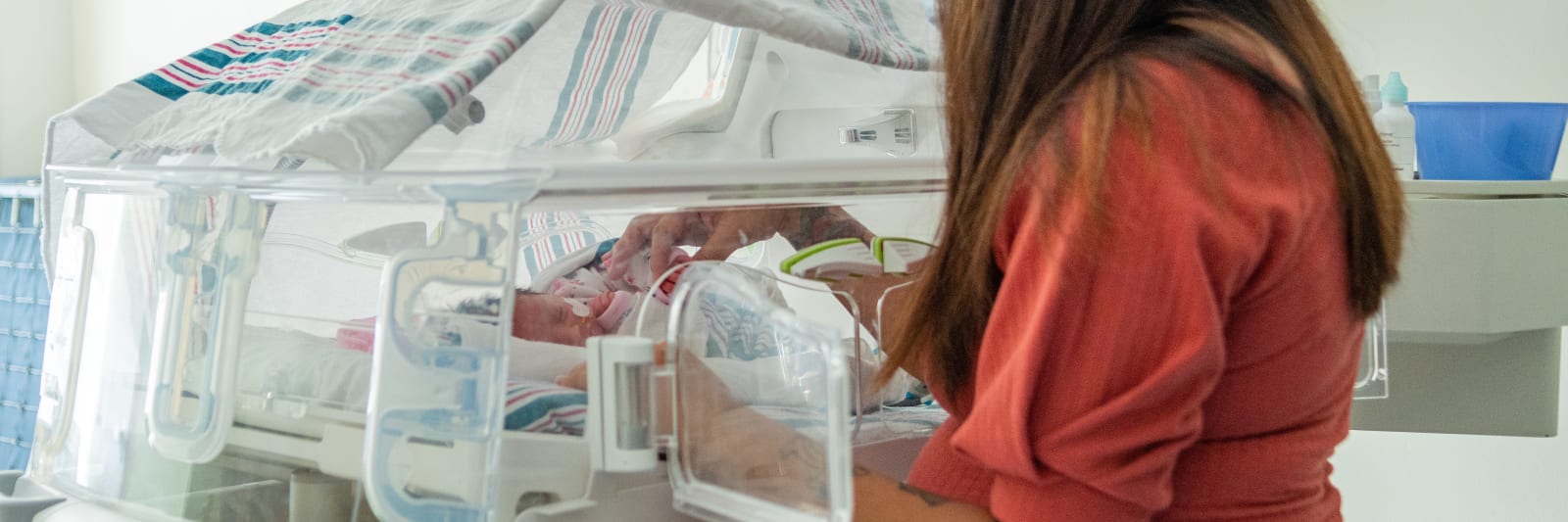 A woman stands next to a NICU incubator and gently places her hands on the baby inside