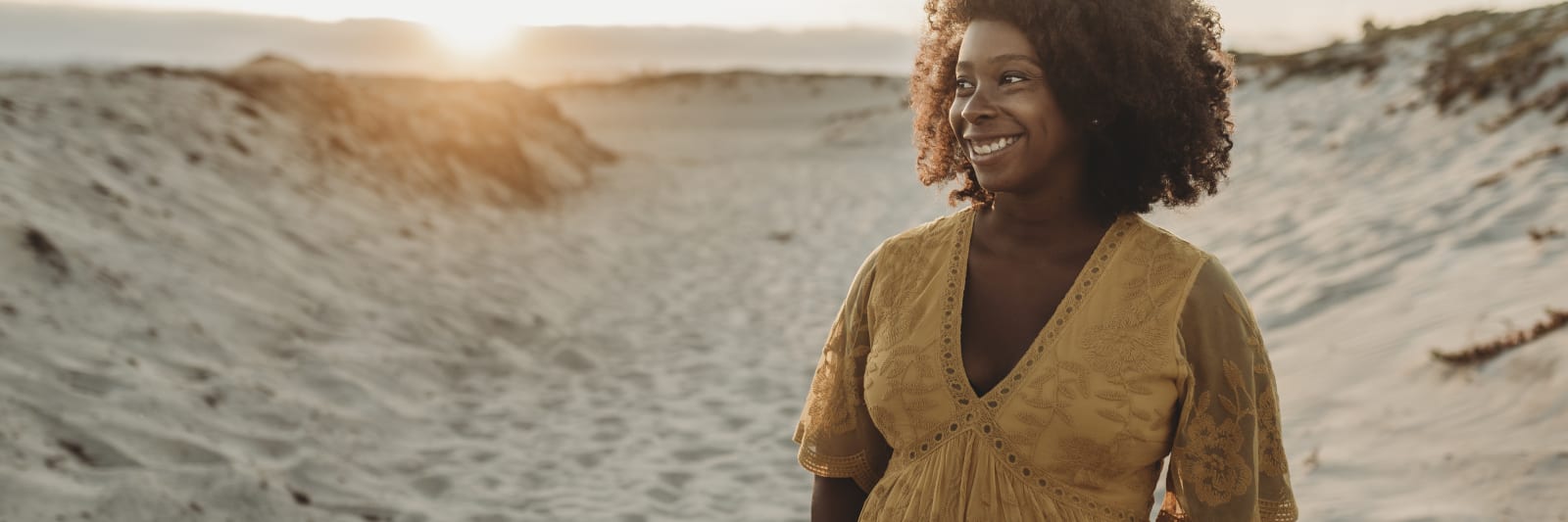 a pregnant woman looks happy cradling her baby bump while she stands in the midst of sand dunes at the oceanfront.