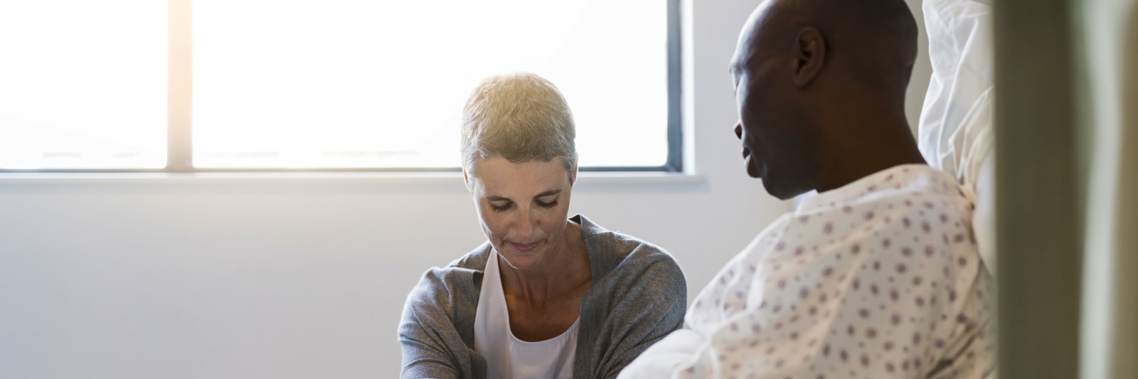 Woman sitting at the bedside of a male patient wearing a hospital gown. They are in brightly lit hospital room.