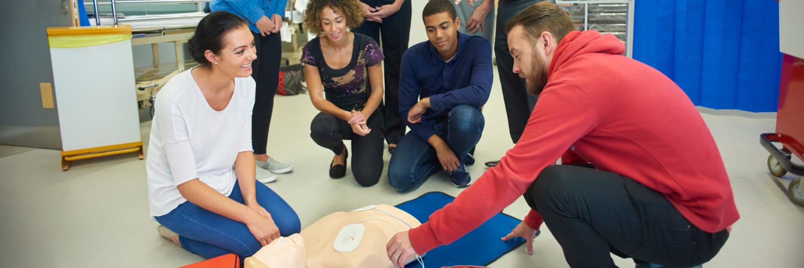 a mixed age group listen to teacher as he shows the procedure involved to resuscitate using a defibrillator