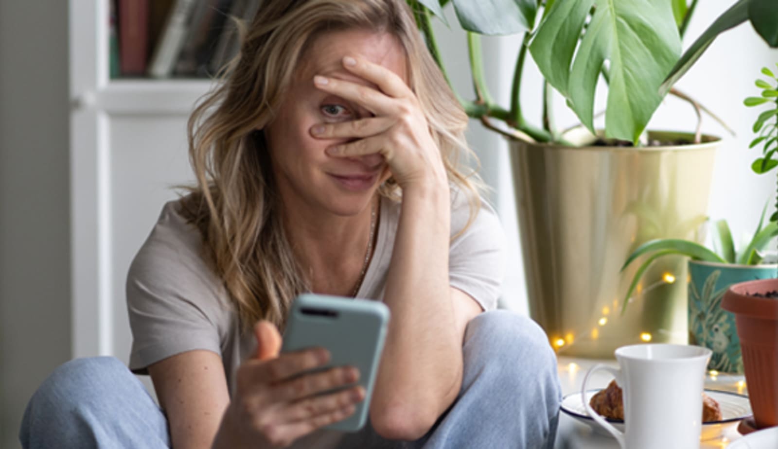 Caucasian woman sitting cross-legged on the floor with her hand over her face while looking at her iPhone. 