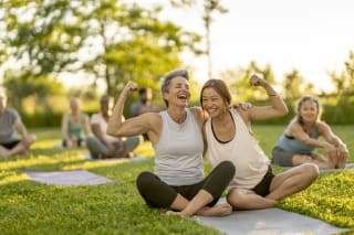 Two laughing woman sit on a yoga mat outside as they lean in closely for a hug and flex their muscles.