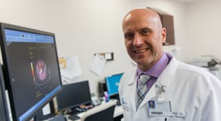 Dr. Edward Gorak smiling at the camera while wearing his doctor lab coat in his workspace with a computer screen.