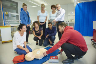 a mixed age group listen to their instructor as he shows the procedure involved to resuscitate using a defibrillator