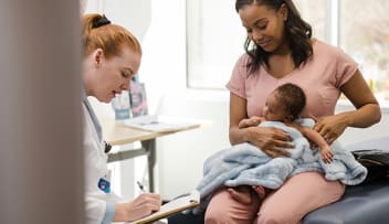 Female doctor updates a newborn's medical record as he and his mother sit on the exam table