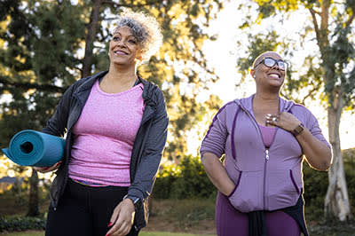 two women in exercise clothing walking outdoors, one is carrying a yoga mat