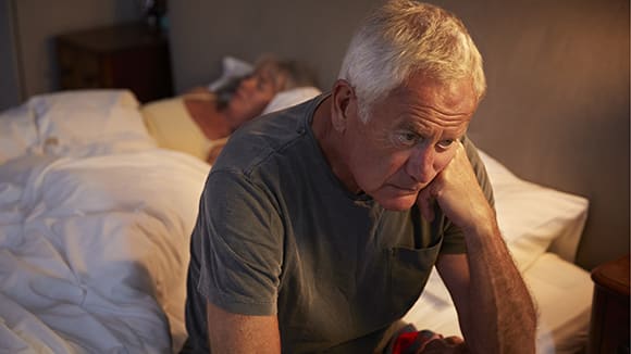 worried looking senior male sitting on the edge of his bed while wife sleeps in the background