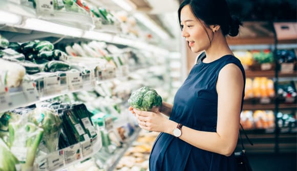 pregnant woman shopping at grocery produce department holding broccoli