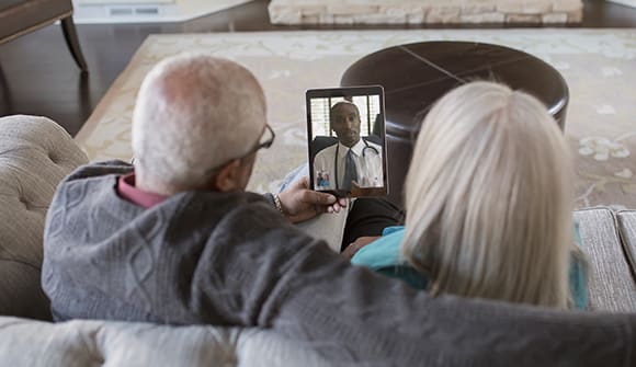 couple sitting on a couch talking to a virtual doctor on their tablet