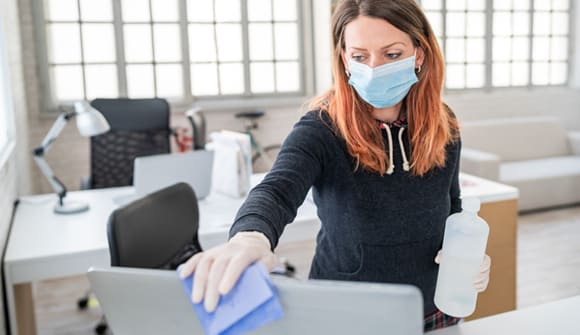 woman wearing a mask and gloves to clean a computer in her office