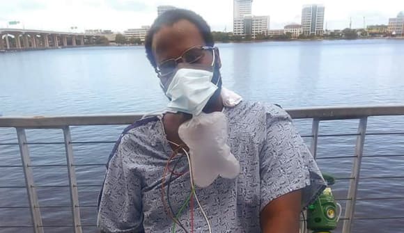 masked man outside in front of the river hooked up to many medical devices