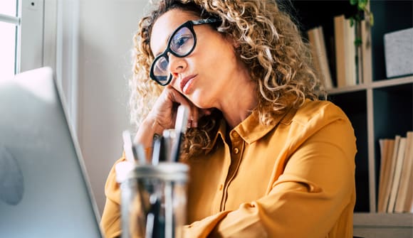 tired looking woman in glasses resting her cheek on her hand as she stares at a computer screen in a home office