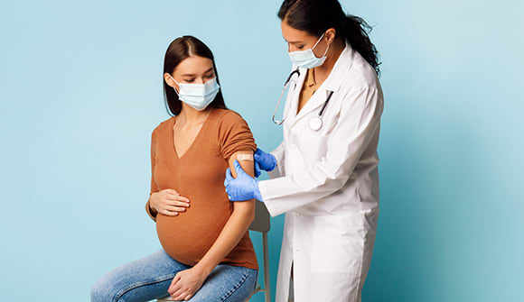 Masked nurse vaccinating pregnant female patient and sticking bandage on her arm