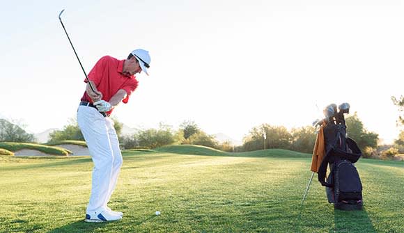 Image of a man playing golf out on the green. The man is wearing a red shirt with white pants and is practicing his golf swing in the early morning hours. 