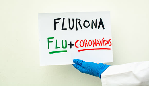 hand in lab coat and blue surgical glove holding a sign with the text fluron, coranavirus + flu, covid-19