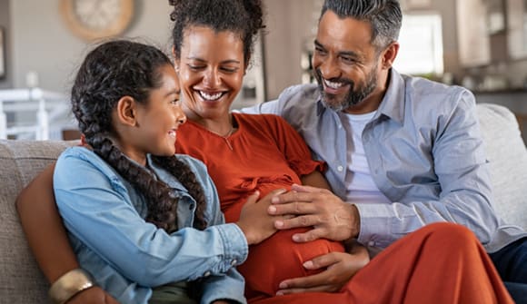 pregnant mother sitting on the couch with partner and daughter feeling her bump