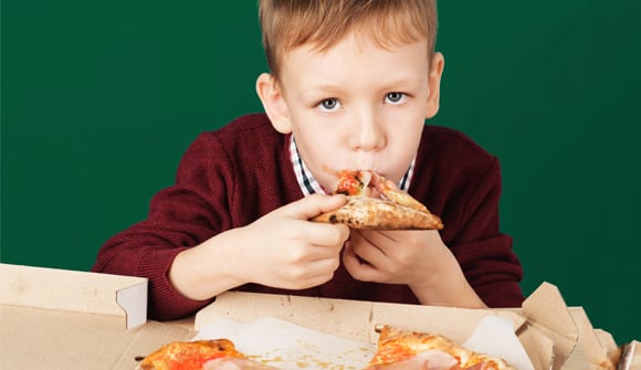 A young boy is staring into the camera as he shoves a cheesy slice of pizza into his mouth