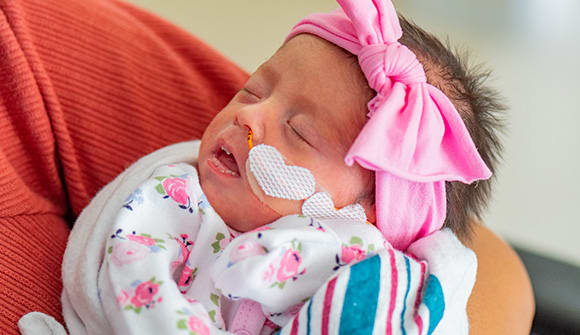 Photo of a premature baby wearign an oversized pink bow wrapped in a pink and white blanket. 