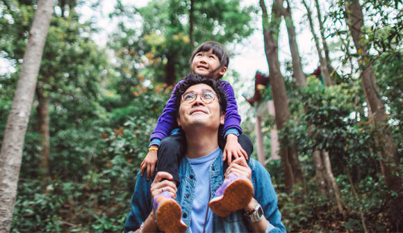 A photo of a young girl sitting on her father's shoulders as they walk through a wooded area during the day. 