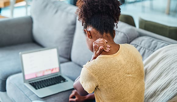 A young woman experiencing neck pain while using a laptop on the sofa at home