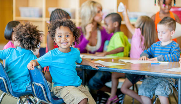 young preschool age child smiles at the camera while sitting at a table with classmates
