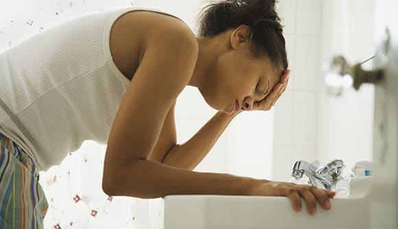 Image of a woman bent over the sink clutching her head with her eyes closed. 