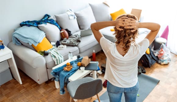 Lady standing with her arms behind her head with her back to the camera facing clutter on her floor at sofa in the background.