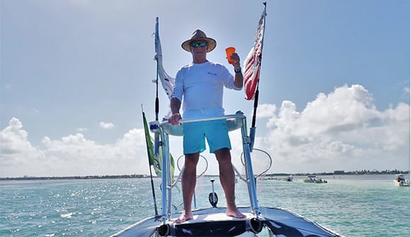 Tom Dodson standing on boat with water and sky in background. An American flag blows behind him. 
