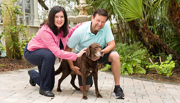 Man in pink jacket smiles on the left. Her fiance is on the right. They are crouched down around their cute chocolate lab.