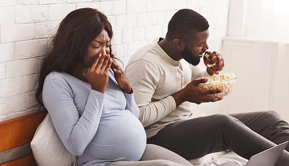 pregnant women with hands to her nose experiencing sensitivity of smell to man eating popcorn